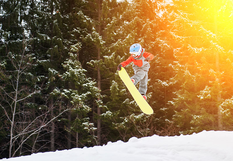 A snowboarder in an orange jacket and blue helmet executes a jump on a snowy slope at Elk Mountain in Union Dale, PA, with a forest of snow-covered trees in the background.