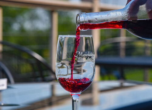 A bottle of red wine is being poured into a Maiolatesi Wine Cellars glass on an outdoor patio table.