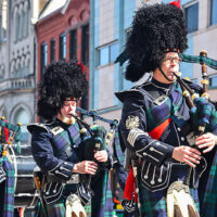 A group of bagpipers in traditional Scottish attire, including kilts and feathered bonnets, are playing during Scranton's St. Patrick's Day Parade.