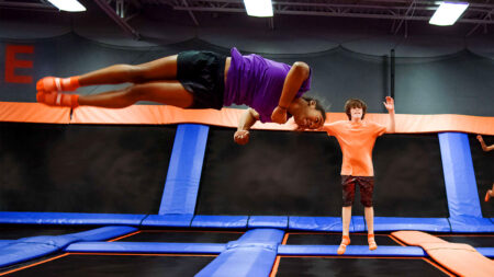 A girl in a purple shirt and orange grip socks does a backflip on a trampoline at Sky Zone Trampoline Park, Pittston, a popular indoor activity for kids, while a boy in an orange shirt jumps nearby.