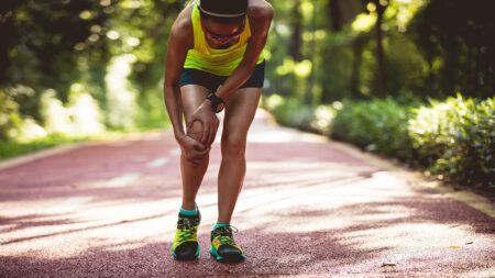 A woman on a running trail stops mid-run and holds her leg in pain.