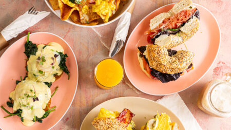 A flat lay of brunch options from The Canning House, Forty Fort, Pa, of four plates of food and a glass of orange juice on a table. The plates contain eggs benedict, a bagel sandwich, breakfast nachos, and another bagel sandwich.