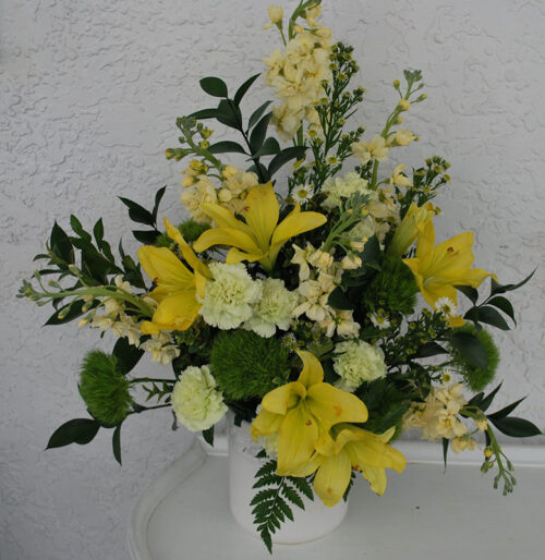 Elegant floral arrangement of yellow lilies, white flowers, and lush greenery displayed in a white ceramic vase against a neutral background.