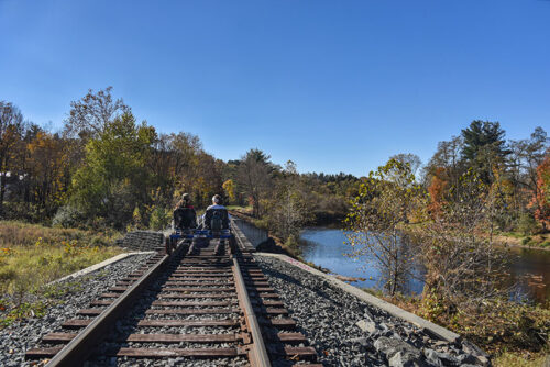 Two people ride a rail bike along the river with Pennsylvania Rail Bike in Hawley, PA.