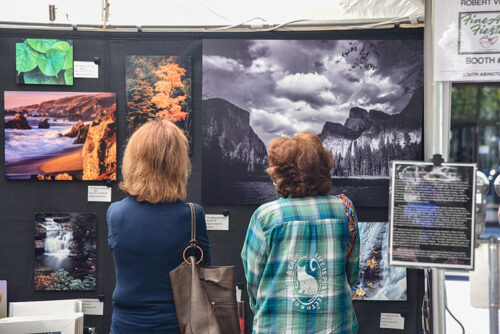Two women are viewing photographs at The Fine Arts Fiesta in Wilkes-Barre, PA. The photos on display feature landscapes, such as a beach, mountains and forests. The women are standing in front of the booth with their backs to the camera.