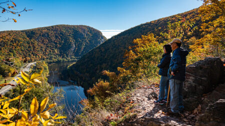 A couple standing on a rocky cliff overlooking the Delaware Water Gap National Recreation Area in autumn, with the Delaware River winding through the valley below and colorful foliage covering the mountains. 