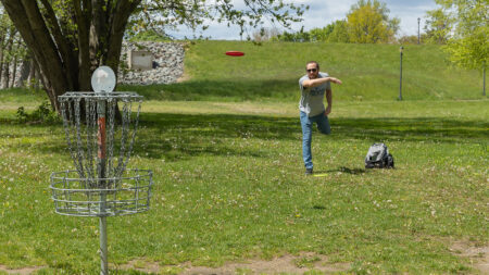 A man in a short sleeve shirt and jeans throwing a frisbee while playing disc golf at Nesbitt Park in Kingston, PA.