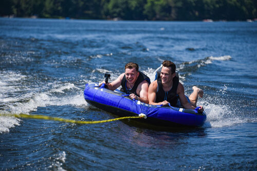 Two smiling young men in life jackets are being towed on a blue and black inflatable tube behind a boat on Lake Wallenpaupack. One man is holding a GoPro camera.
