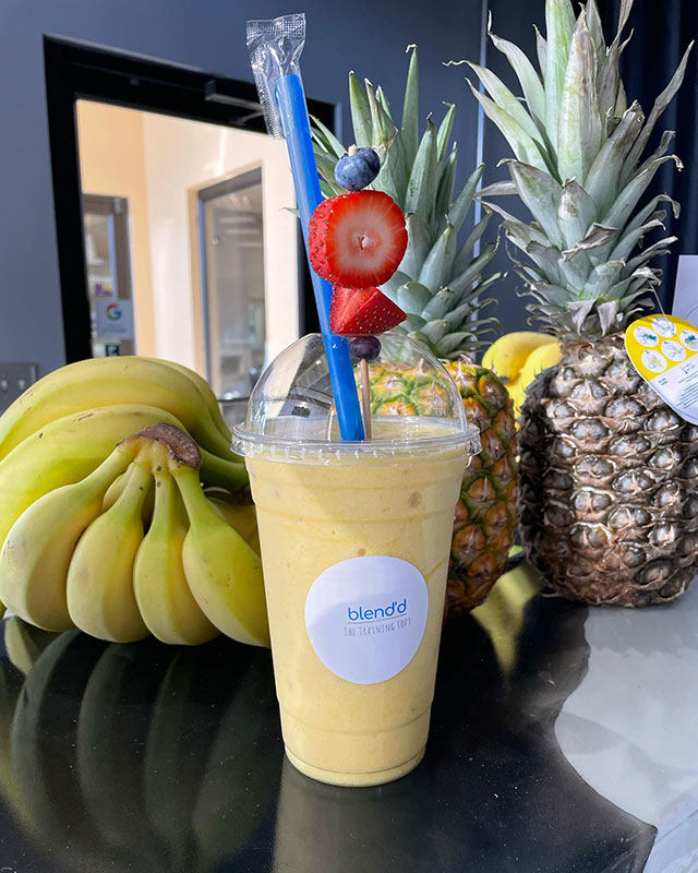 A yellow smoothie topped with strawberries and blueberries with fruit in the background from Blend'd Smoothie Bar in Clarks Summit, PA.