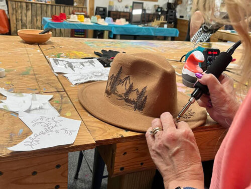 A close-up of a person's hands using a wood-burning tool to etch a mountain and tree design onto a light brown felt hat at the Back Mountain Makery in Dallas, Pennsylvania.