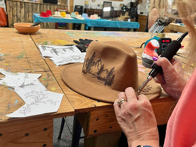 A close-up of a person's hands using a wood-burning tool to etch a mountain and tree design onto a light brown felt hat at the Back Mountain Makery in Dallas, Pennsylvania.