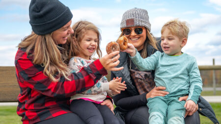 A woman holding a girl on her lap sits next to another woman holding a boy, as they toast their apple cider donuts, a favorite fall treat at Roba Family Farms in North Abington Twp., PA.