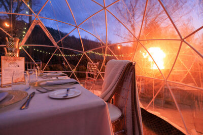 A dining igloo with a set table sits next to a roaring fire pit at twilight at The Beaumont Inn, Dallas, PA. The igloo is strung with fairy lights and a blanket is draped over one of the chairs.