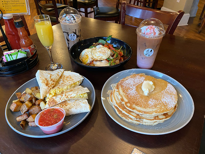 A wooden table at Taylor Deli & Café in Taylor, Pa, holds a variety of breakfast foods. There are quesadillas, potatoes and a small bowl of salsa on one plate, a bowl of eggs and vegetables, and a stack of pancakes another plate. There is a glass of orange juice and two milkshakes on the table as well.