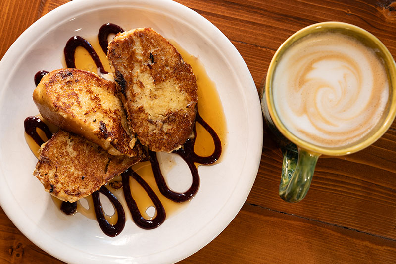 A plate of French toast at Sugar and Loaf Bakery in Drums, Pa, topped with syrup and a swirl of chocolate sauce sits next to a mug of latte on a wooden table.