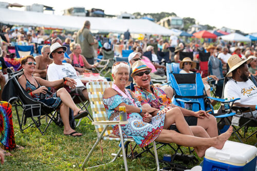 An older couple on lawn chairs amongst the crowd in the grass smile at the camera during a performance at the Briggs Farm Blues Festival in Nescopeck, PA.