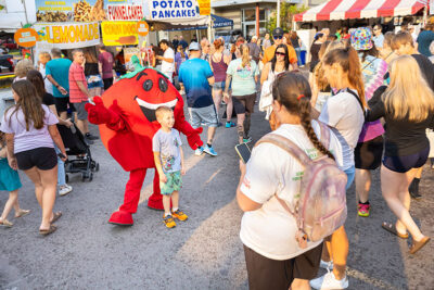 A young boy poses with the Pittston Tomato Festival mascot, a large tomato character, amidst a crowd of people and food stands at the annual festival in Pittston, PA.