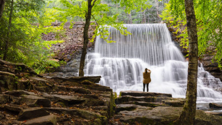 A lone woman stands on the rocks framed between two trees and photographs a cascading waterfall along the Shades of Death trail at Hickory Run State Park in White Haven, PA.