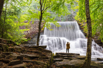 A lone woman stands on the rocks framed between two trees and photographs a cascading waterfall along the Shades of Death trail at Hickory Run State Park in White Haven, PA.