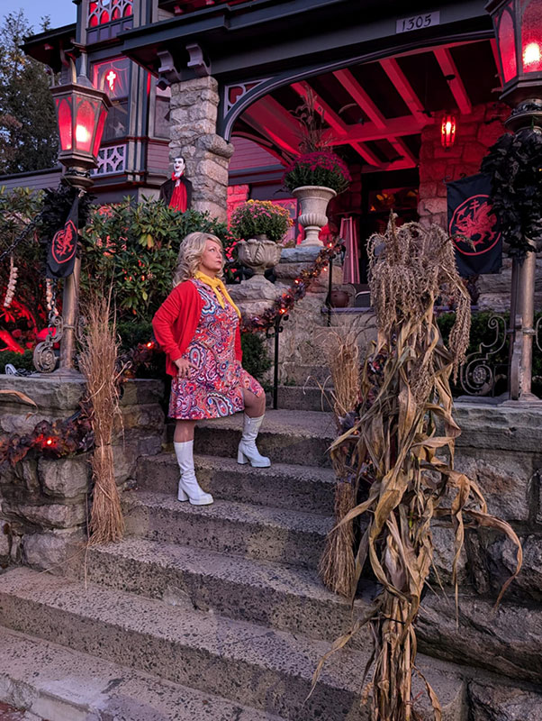 A woman in a retro outfit poses on steps of a Halloween-decorated house with red lighting and spooky decor.