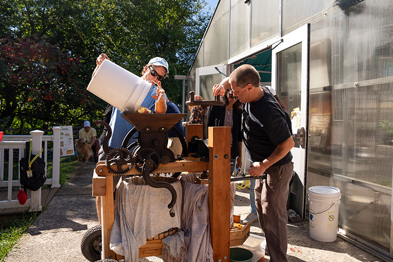 Two men operate a traditional apple press outdoors next to a greenhouse at The Greenhouse Project, Scranton, PA, with one pouring apples into the top while the other cranks the mechanism.