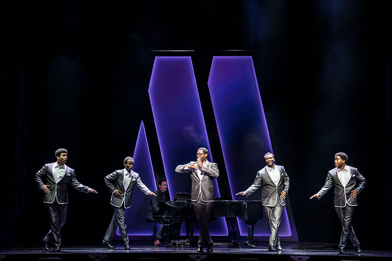 Five men portraying The Temptation sing on stage alongside a piano. 