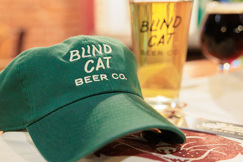 A green baseball hat with the words: Blind Cat Beer Co. in front of two pints of beer at Blind Cat Beer Co. in Pittston, PA.