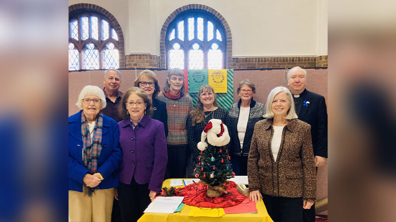 A group of adults stands smiling behind a decorated holiday table with a small snowman centerpiece inside a room with arched windows and festive banners.