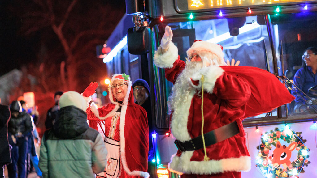 Santa and Mrs. Claus greet bundled-up families with cheerful waves as a festively lit bus glows behind them at the CAN DO Community Christmas and City Hall Tree Lighting in Hazleton, PA.