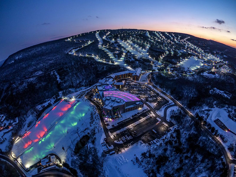 The sun sets behind an illuminated Camelback Mountain. A panoramic view shows the lighted ski trails, the resort grounds and the red and green lights reflecting on the snow tube lanes at Camelback Resort in Tannersville, PA.