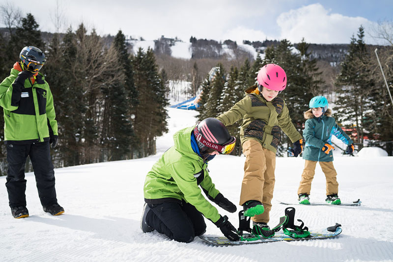 Skin instructors in green jackets help a young girl in a pink helmet strap into her snowboard on the beginner slope just before a lesson.