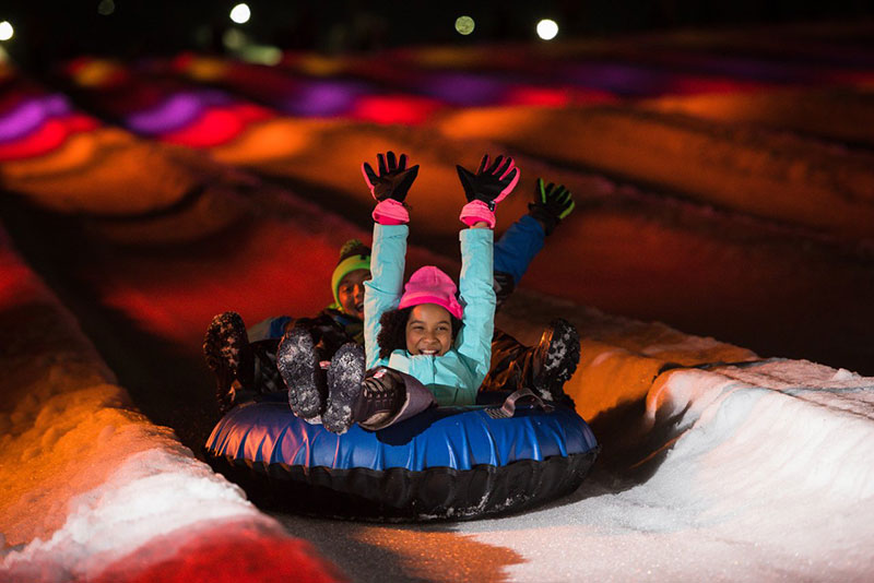 Two children wave their hands and smile while enjoying a ride in a snow tube down the illuminated lanes at Camelback Resort in Tannersville, PA.