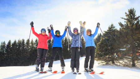 Four women skiers at the top of a ski slope smiling at the camera with their hands raised in the air at Elk Mountain in Union Dale, PA.