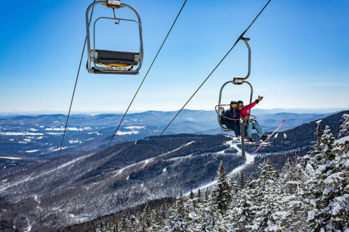 Two people up on a chair lift, riding to the top of the ski slope, waving at the camera below them at Sugarbush Resort in Vermont.