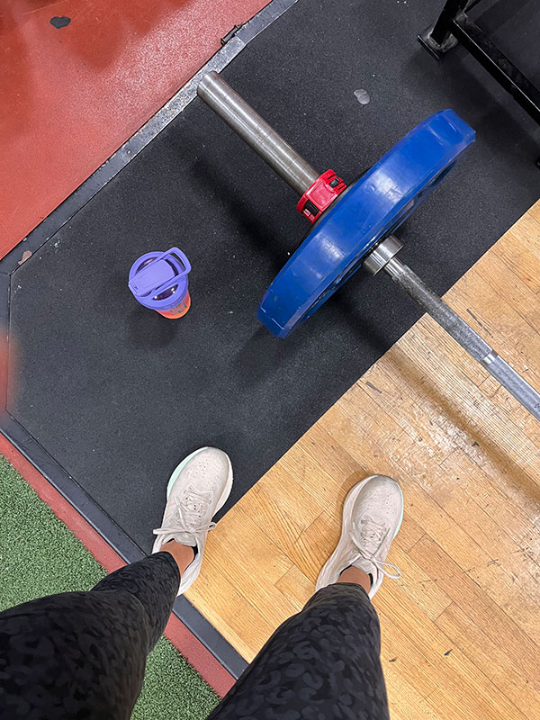 A person standing on a gym floor next to a barbell with a blue weight plate and a water bottle.