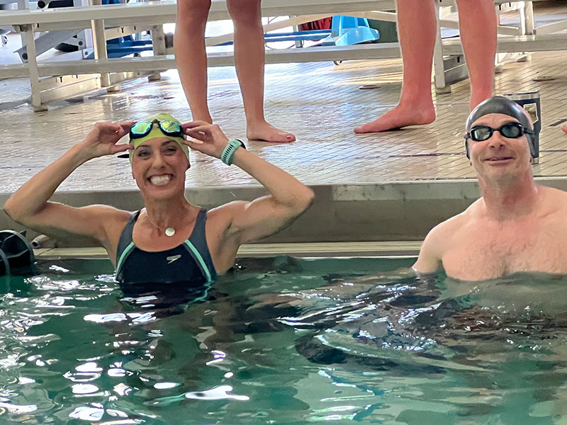 A woman and a man in an indoor pool are smiling at the camera. The woman is adjusting the goggles on her head.