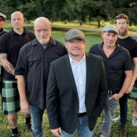 A photo of the Celtic rock band, Kilmaine Saints, performing at Mauch Chunk Opera House, featuring seven members in casual attire, some wearing kilts and caps, posing outdoors against a backdrop of trees.