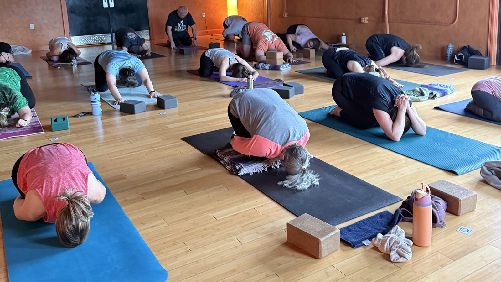 A yoga class is in session, with multiple participants in child's pose on their mats in a studio with wood floors.