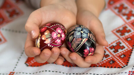 A close up shot of a person's hands holding two intricately decorated easter eggs. The egg on the left is maroon with pink and orange painted flowers while the egg on the right is black with grey and red painted flowers.