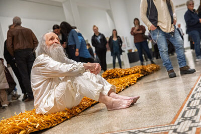 A man dressed all in white and sitting on cornstalk matting smiles in the distance in an art gallery full of people at the Everhart Museum in Scranton, PA.