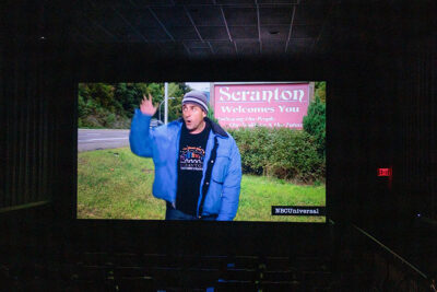 A still shot of the character Michael Scott appears on a movie theater screen as he stands in front of a sign reading 