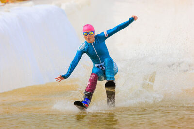 A female skier, dressed in a blue skinsuit and a pink swim cap, splashes across a pond at Camelback Resort's 20th Pond Skim event in Tannersville, PA.