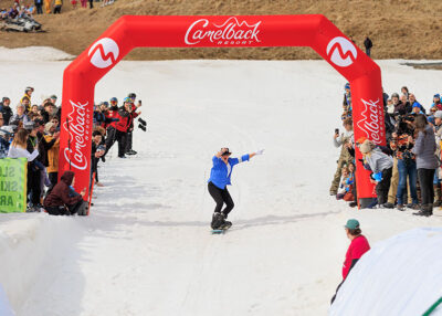 A female snowboarder dressed as Michael Jackson rides under a red, inflatable arch, while spectators cheer her on at Camelback Resort's 20th Pond Skim event in Tannersville, PA.