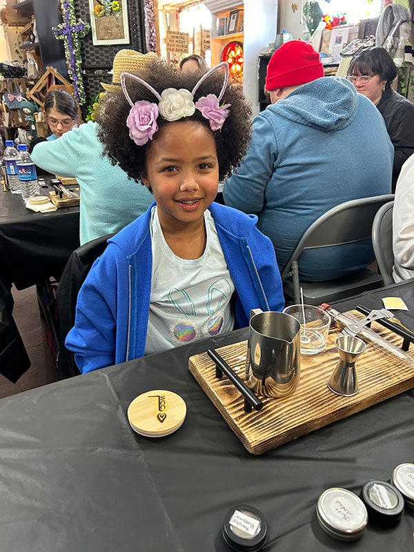 A young girl with pink flowers in her hair begins work at a table during a candle making class at the Local Emporium Co. in Tunkhannock, PA.