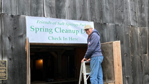 An older man standing on a ladder in front of a banner for the Spring Clean Up Day hanging in a barn doorway at Salt Springs Park.