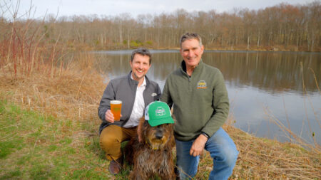Two men pose with a shaggy brown dog in a hat next to the lake at Curran Brewing in Madison Twp., PA.