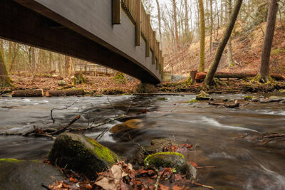 A wooden bridge crosses over a forested stream in early spring at Marshalls Falls Park in East Stroudsburg, PA.
