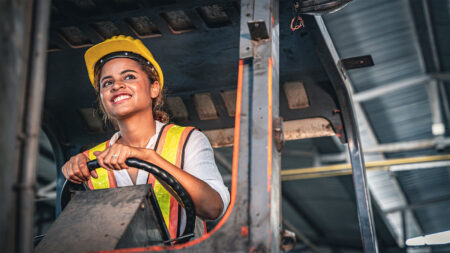 A young woman in a hard hat and safety vest drives a forklift.