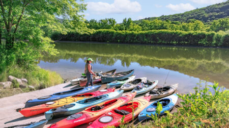 A woman stands near a collection of colorful kayaks on the bank of a river in Northeastern Pennsylvania.