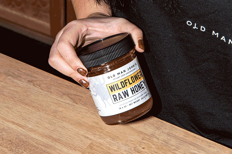 Woman's hand holding a car of Wildflower Raw Honey over the counter at Old Man John’s Coffee and Honey House in Luzerne, PA.
